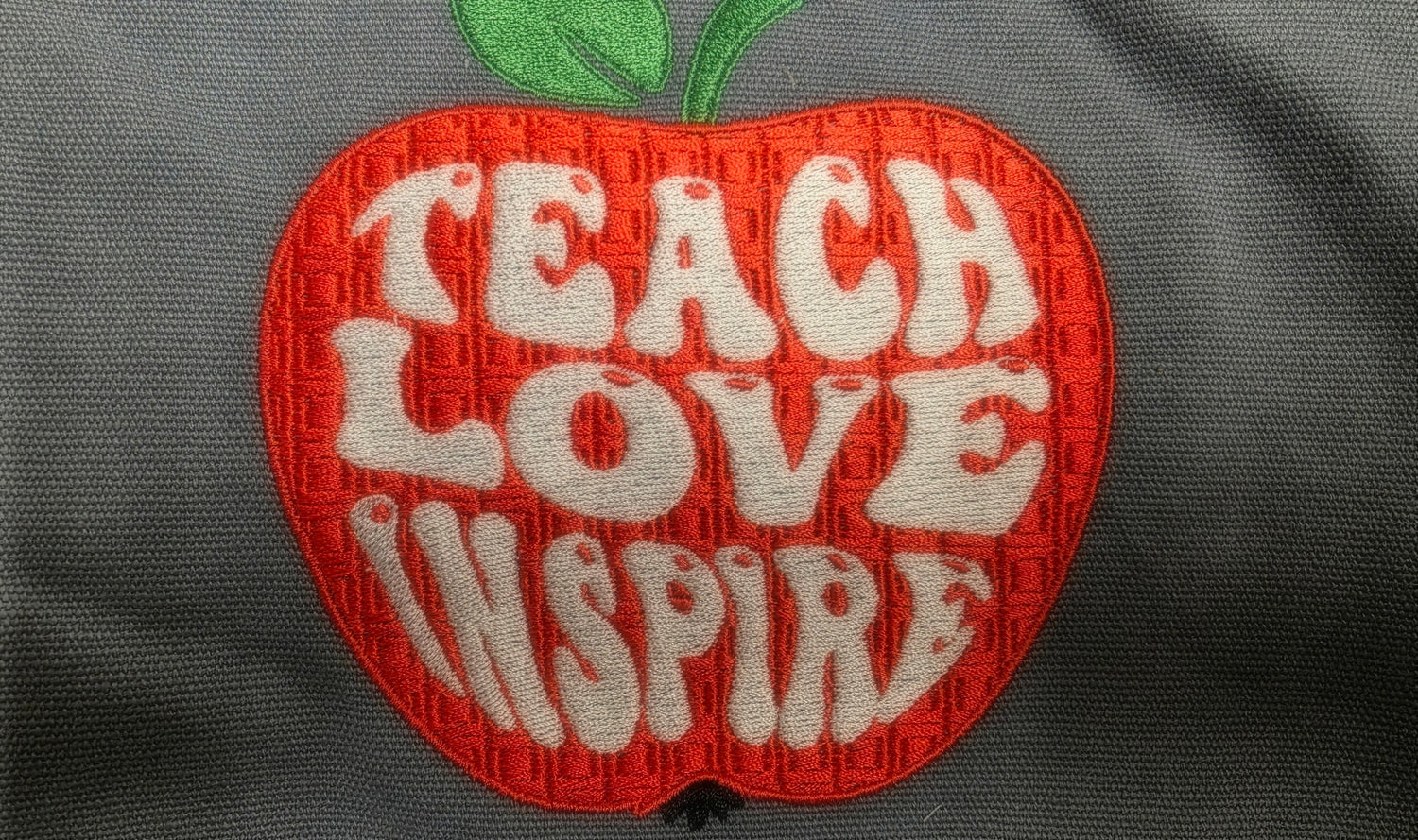 Embroidered patch in the shape of an apple with 'TEACH LOVE INSPIRE' text on a dark background